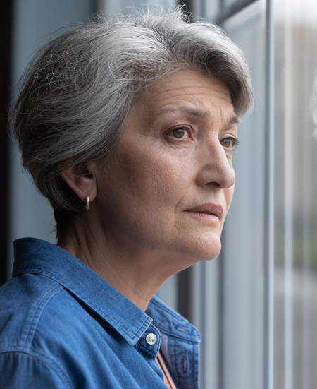 An older woman looks out a window, melancholic.